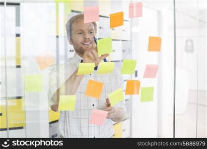 Young businessman reading sticky paper on glass wall at creative office