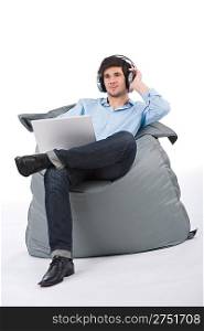 Young businessman relax with music and laptop sitting on bean bag on white background