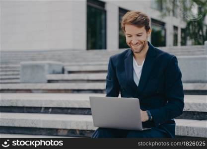 Young businessman sits on steps uses laptop organizes meeting online with investors has happy expression uses free internet poses outdoor dressed formally. Corporate director analyzes web information. Young businessman sits on steps uses laptop organizes meeting online with investors