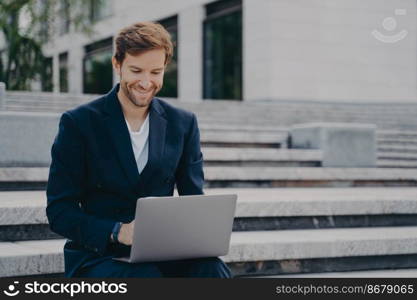 Young businessman sits on steps uses laptop organizes meeting online with investors has happy expression uses free internet poses outdoor dressed formally. Corporate director analyzes web information. Young businessman sits on steps uses laptop organizes meeting online with investors