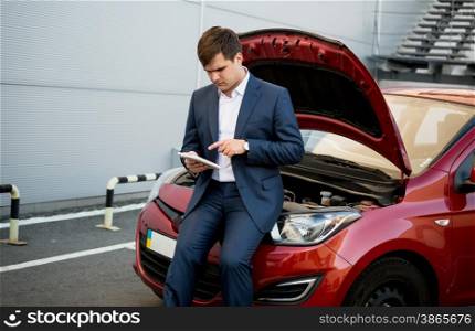 Young businessman sitting on hood of broken car and searching how to fix it on tablet