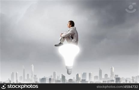 Young businessman sitting on light bulb top and thinking something over. Waiting for inspiration