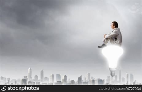 Young businessman sitting on light bulb top and thinking something over. Waiting for inspiration