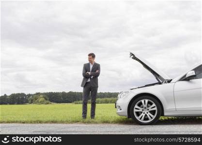 Young businessman standing arms crossed by broken down car at countryside