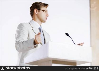 Young businessman standing on stage and speaking to microphone . Speaker at stage
