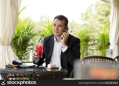 Young businessman talking on a cell phone and drinking a cocktail sitting in a summer restaurant