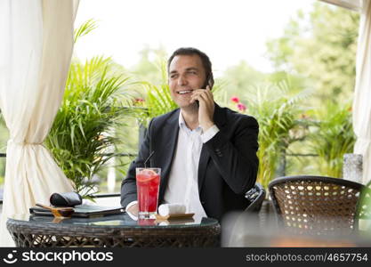 Young businessman talking on a cell phone and drinking a cocktail sitting in a summer restaurant