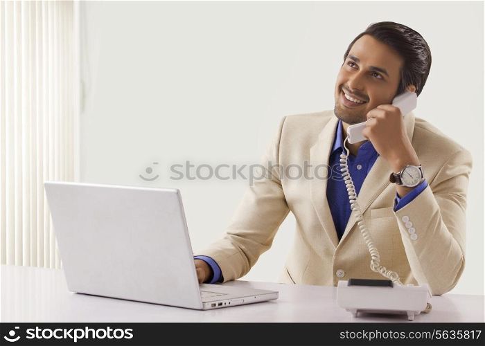 Young businessman talking on telephone while using laptop at office desk