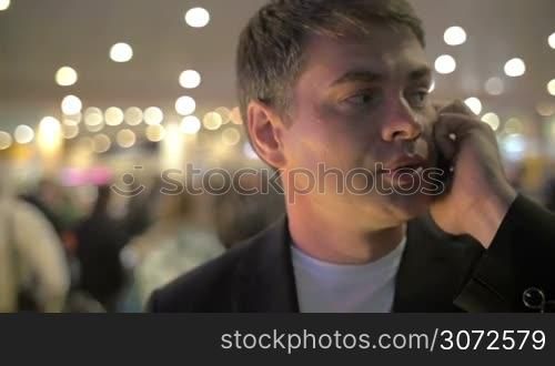Young businessman talking on the phone while waiting for the flight at the airport. Defocused crowd and electric lights in background