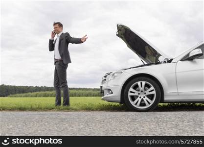 Young businessman using cell phone by broken-down car at countryside