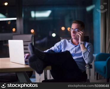 Young businessman using mobile phone while working on laptop at night in dark office.