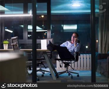 Young businessman using mobile phone while working on laptop at night in dark office.