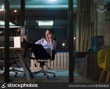 Young businessman using mobile phone while working on laptop at night in dark office.