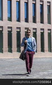 Young businessman walking next to office buildings while holding a shoulder bag outdoors