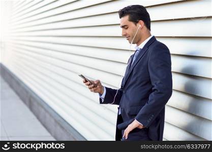 Young businessman wearing blue suit and tie using a smartphone in urban background. Man with formal clothes and headphones in the street.