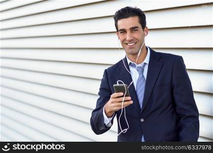 Young businessman wearing blue suit and tie using a smartphone in urban background. Man with formal clothes and headphones in the street. Good looking guy looking at camera with toothy smile.