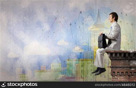 Young businessman with suitcase sitting on roof of building. Businessman on top