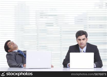 Young businessman working on laptop while colleague sleeping at desk in office
