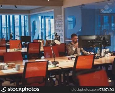 Young businessman working overtime alone at his desk on desktop computer in an open space modern coworking office late at night with city lights glowing in the background. High quality photo. businessman working overtime alone at his desk in an office late at night 