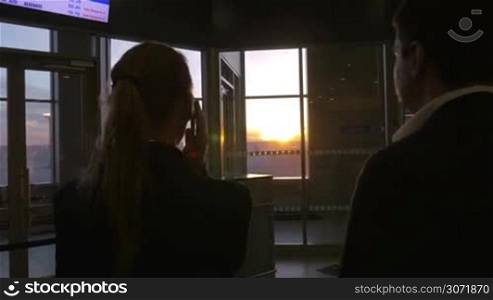 Young businesspeople at the airport terminal. Woman having a phone talk, man looking out the window and enjoying sunset scene