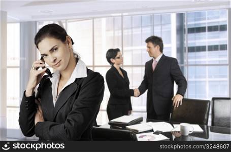 Young businesswoman calls on cell phone while other business people are shaking hands in the background. Daylight, indoor, office.