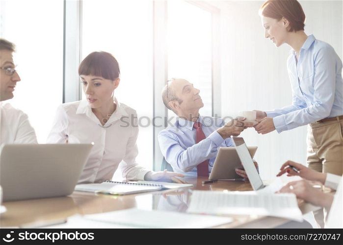 Young businesswoman giving coffee to businessman by colleagues during meeting in boardroom at modern office