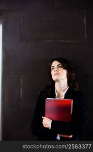 Young businesswoman in dark suit holding red folder daydreaming on office corridor posing for business portrait.