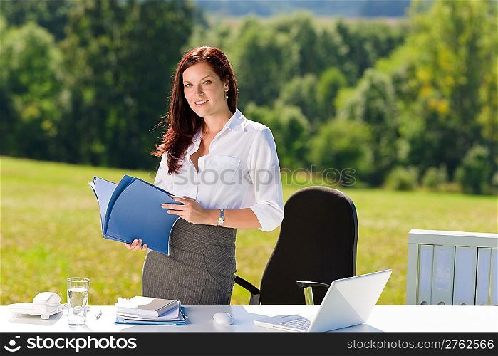 Young businesswoman in sunny nature office holding folders behind table