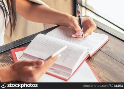 Young businesswoman is writing plans on the notebook while holding smartphone at cafe. Business concept. Young businesswoman is writing plans on the notebook