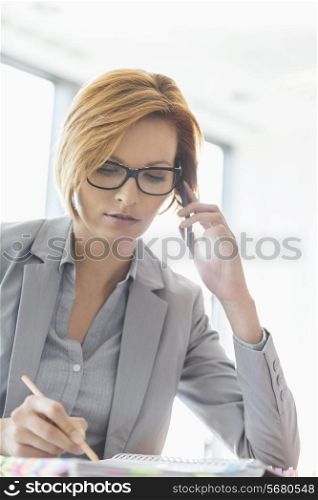 Young businesswoman on call while writing at desk in office