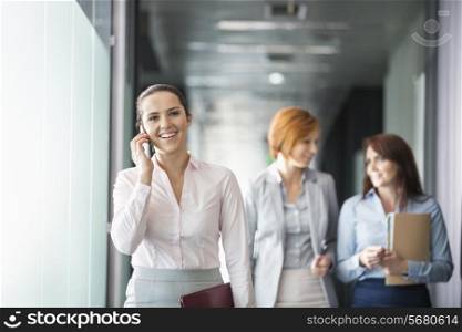Young businesswoman on call with colleagues in background at office corridor