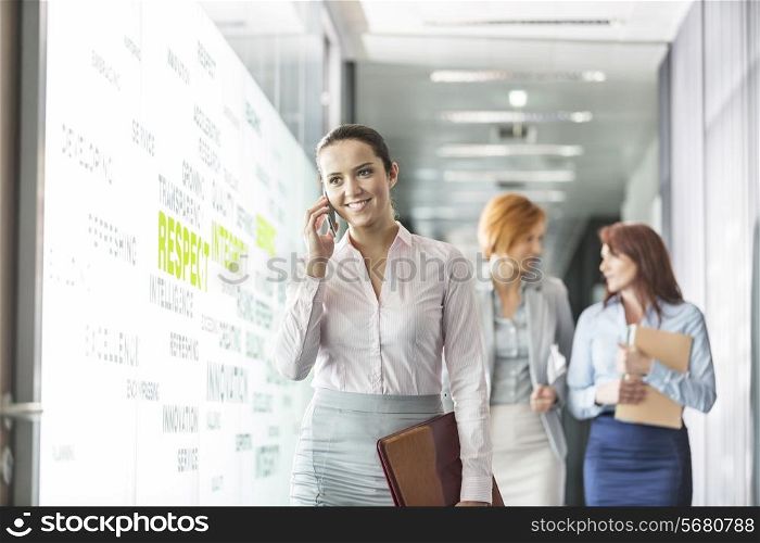 Young businesswoman on call with colleagues in background at office corridor