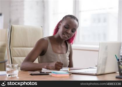 Young businesswoman sitting at workplace and writing numbers in notebook after calculating budget