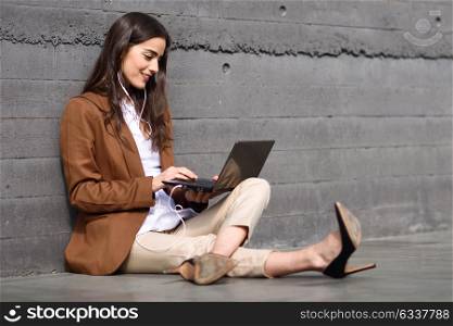 Young businesswoman sitting on floor looking at her laptop computer. Beautiful woman wearing formal wear using earphones.