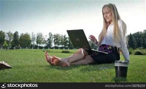 Young businesswoman sitting on green grass in the park and talking with business partners online using a laptop