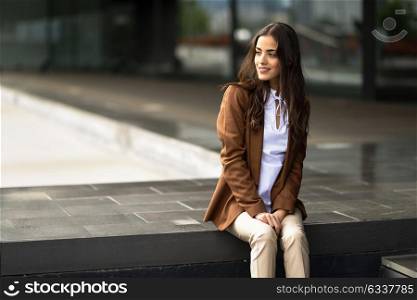 Young businesswoman sitting outside of office building. Beautiful woman wearing formal wear. Young girl with brown jacket and trousers in urban background.
