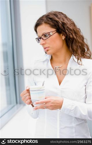 Young businesswoman standing in front of office window, drinking coffee and thinking.