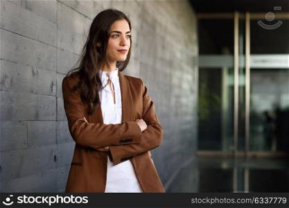 Young businesswoman standing outside of office building. Beautiful woman wearing formal wear. Young girl with brown jacket and trousers.