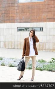Young businesswoman standing outside of office building with briefcase. Beautiful woman wearing formal wear. Young girl with brown jacket and trousers.