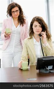 Young businesswoman talking on phone while coworker holding coffee cup.