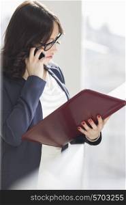 Young businesswoman using cell phone while reading file in office