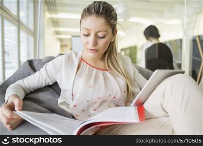 Young businesswoman with digital tablet reading book in office