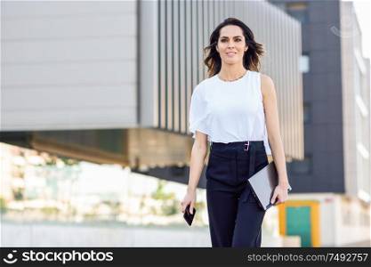 Young businesswoman working with her smart phone and laptop outdoors. Middle-aged woman walking near business building with very careful hair.. Businesswoman working with her smart phone and laptop outdoors.