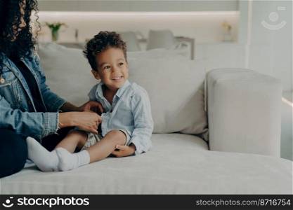 Young caring mother dressing up her little cute son with curly hair, putting on adorable boy shirt and buttoning it, getting ready to walk in park while sitting on sofa at home. Family and childcare. Caring afro american mom dressing up little son while sitting on sofa in living room