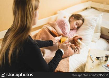 Young caring mother giving hot tea to daughter lying in bed