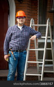 Young carpenter in hard hat posing at metal step ladder