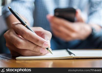 Young casual Business man writing something on notebook and using touchscreen smartphone in the cafe. business, lifestyle, technology concept