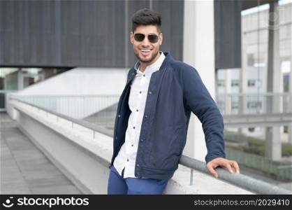 Young casual man looking at camera, wearing sunglasses is standing in modern city downtown. High quality photo.. Young casual man looking at camera, wearing sunglasses is standing in modern city downtown.