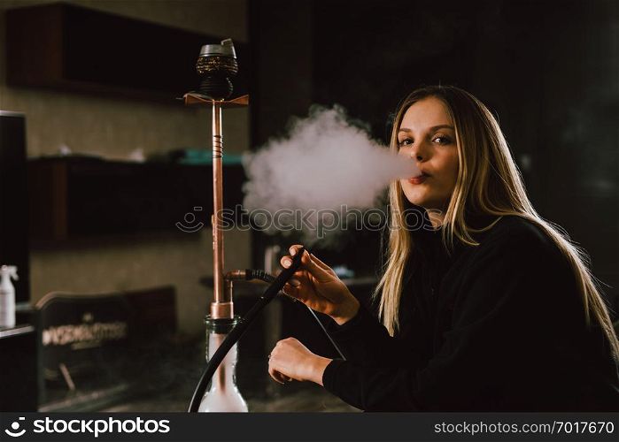 Young caucasian blonde girl smoking hookah at luxury interior with custom v8 car engine table. Leather sofa and beautiful smile
