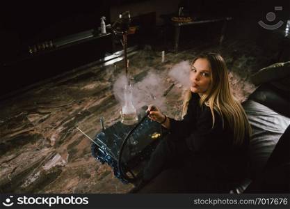 Young caucasian blonde girl smoking hookah at luxury interior with custom v8  car engine table. Leather sofa and beautiful smile